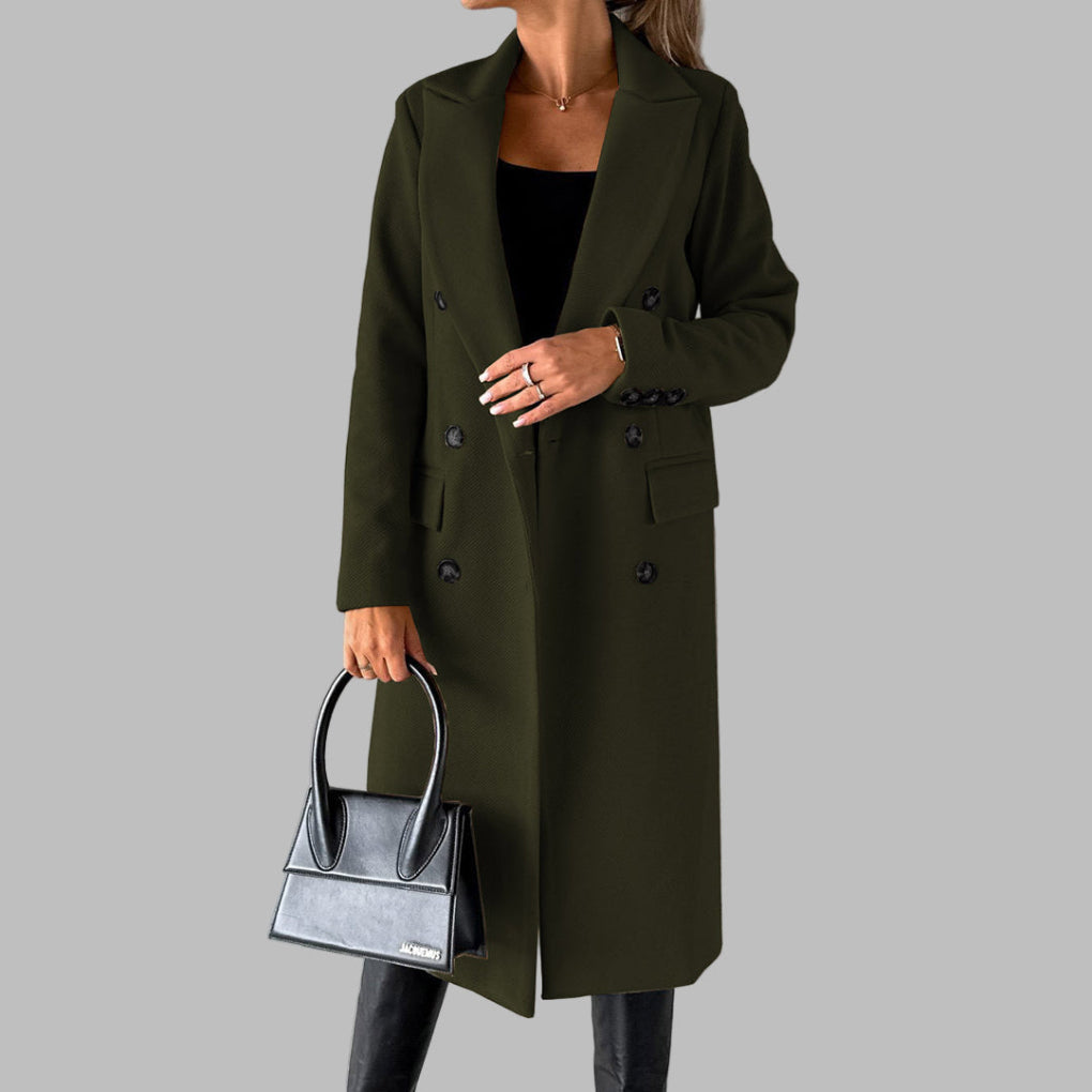 Vrouw in olijfgroene winterjas met dubbele knopen, zwarte handtas en zwarte laarzen tegen een grijze achtergrond. Stijlvolle damesmode.